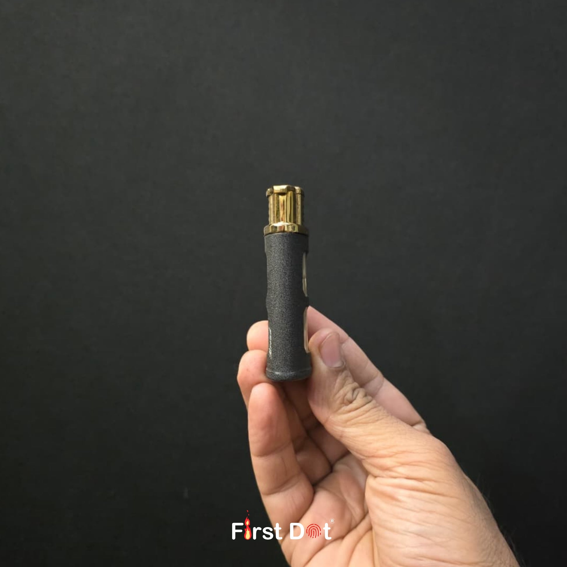 Hand holding a black and gold cylindrical object against a dark background with 'First Dot' branding.