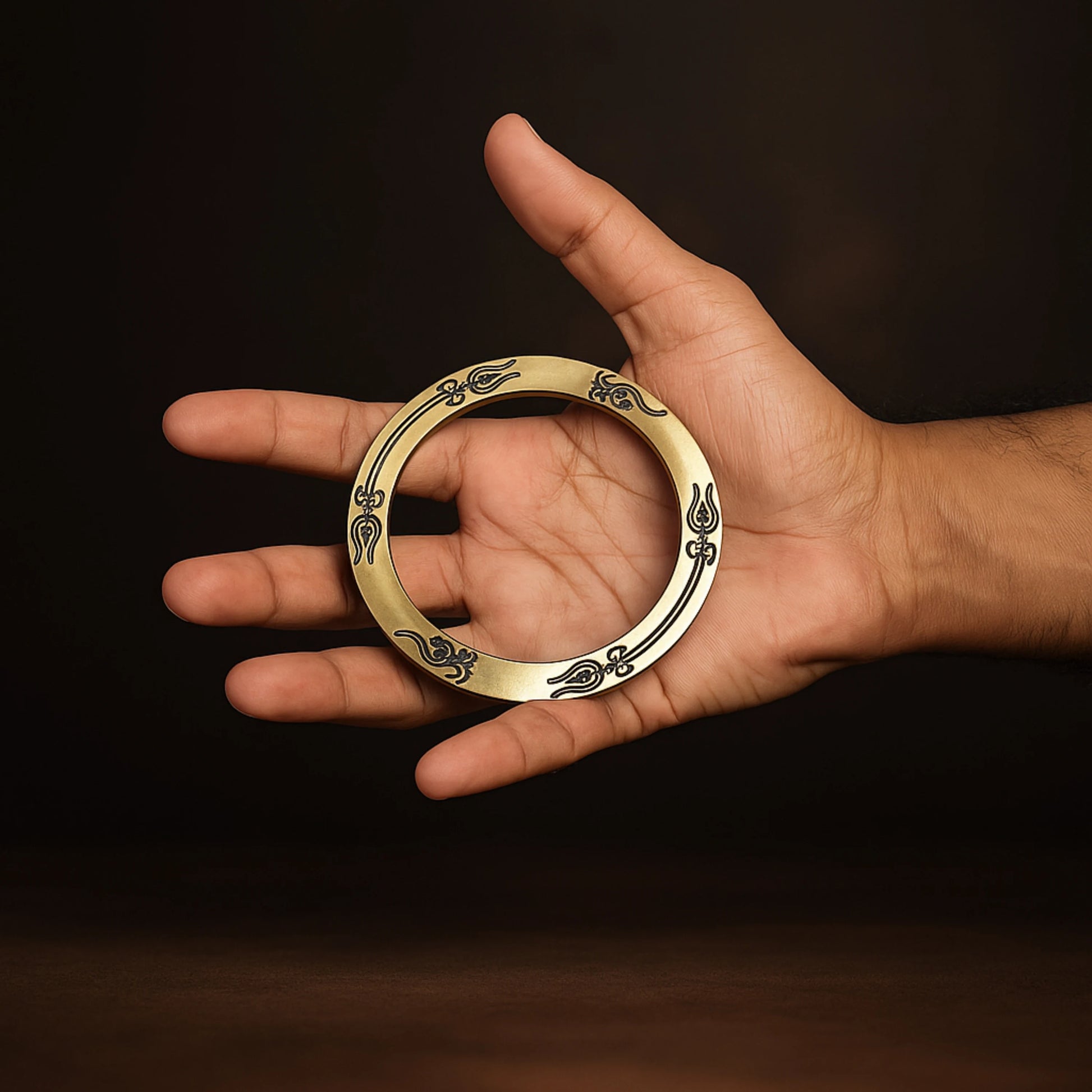 Hand holding a gold ring with intricate designs against a dark background महाकाल Brass Kada