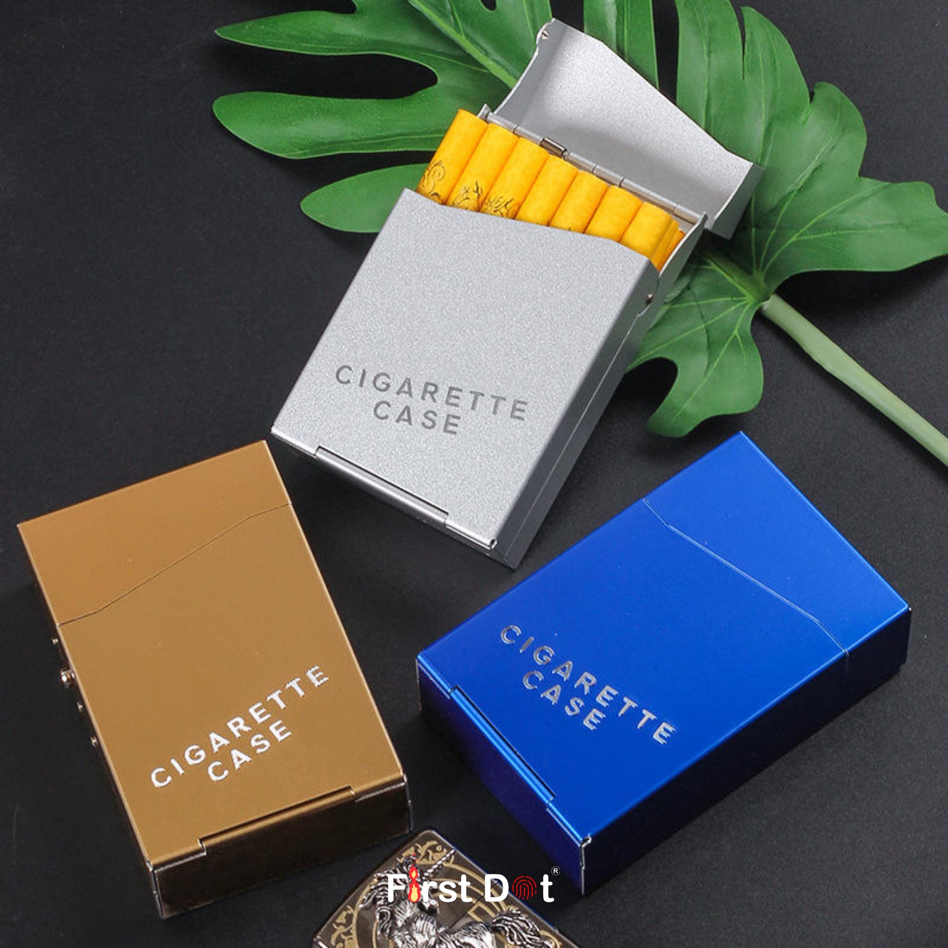 Three cigarette cases in gold, silver, and blue on a dark surface with green leaves.
