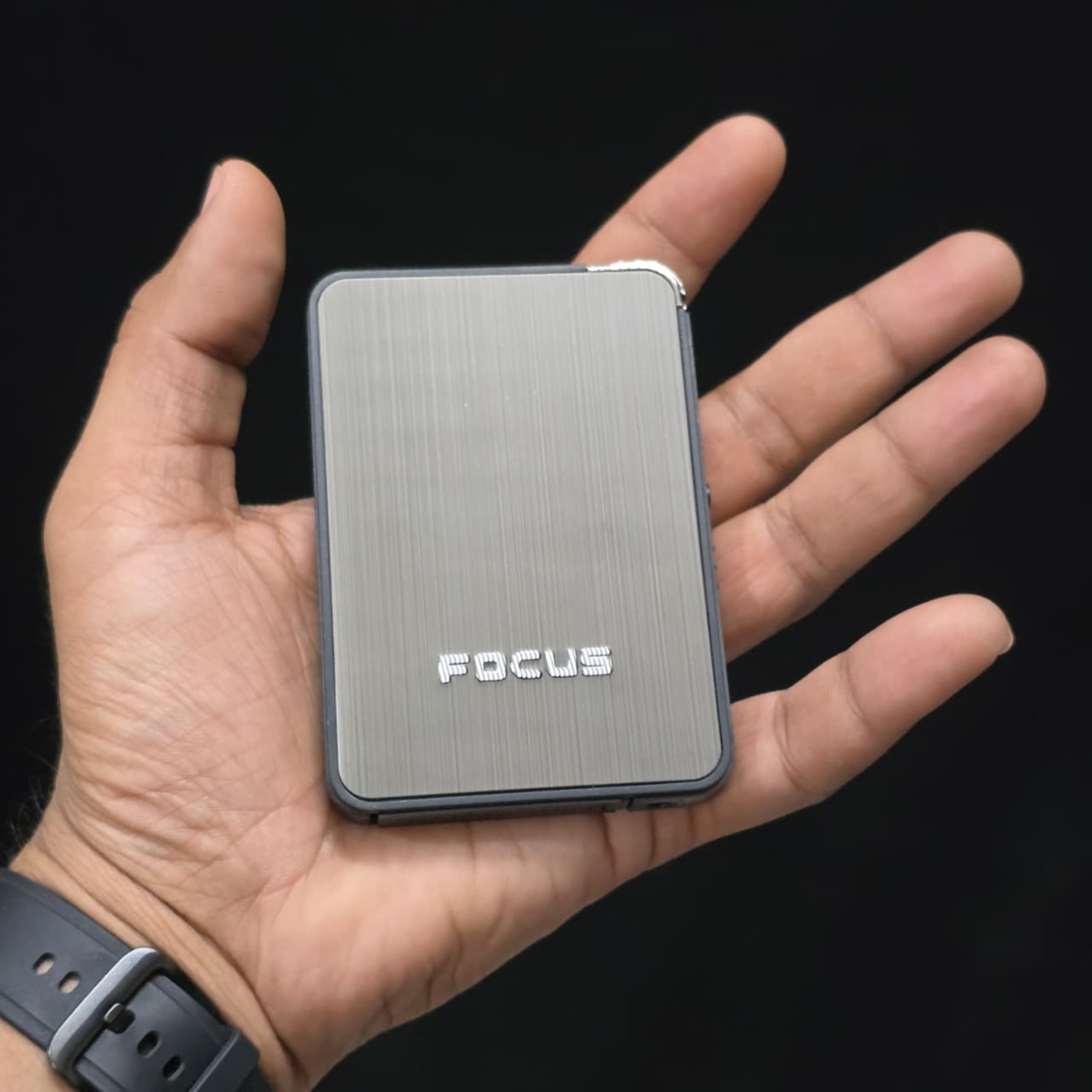 Hand holding a cigarette case with built-in lighter 'FOCUS' branding against a black background