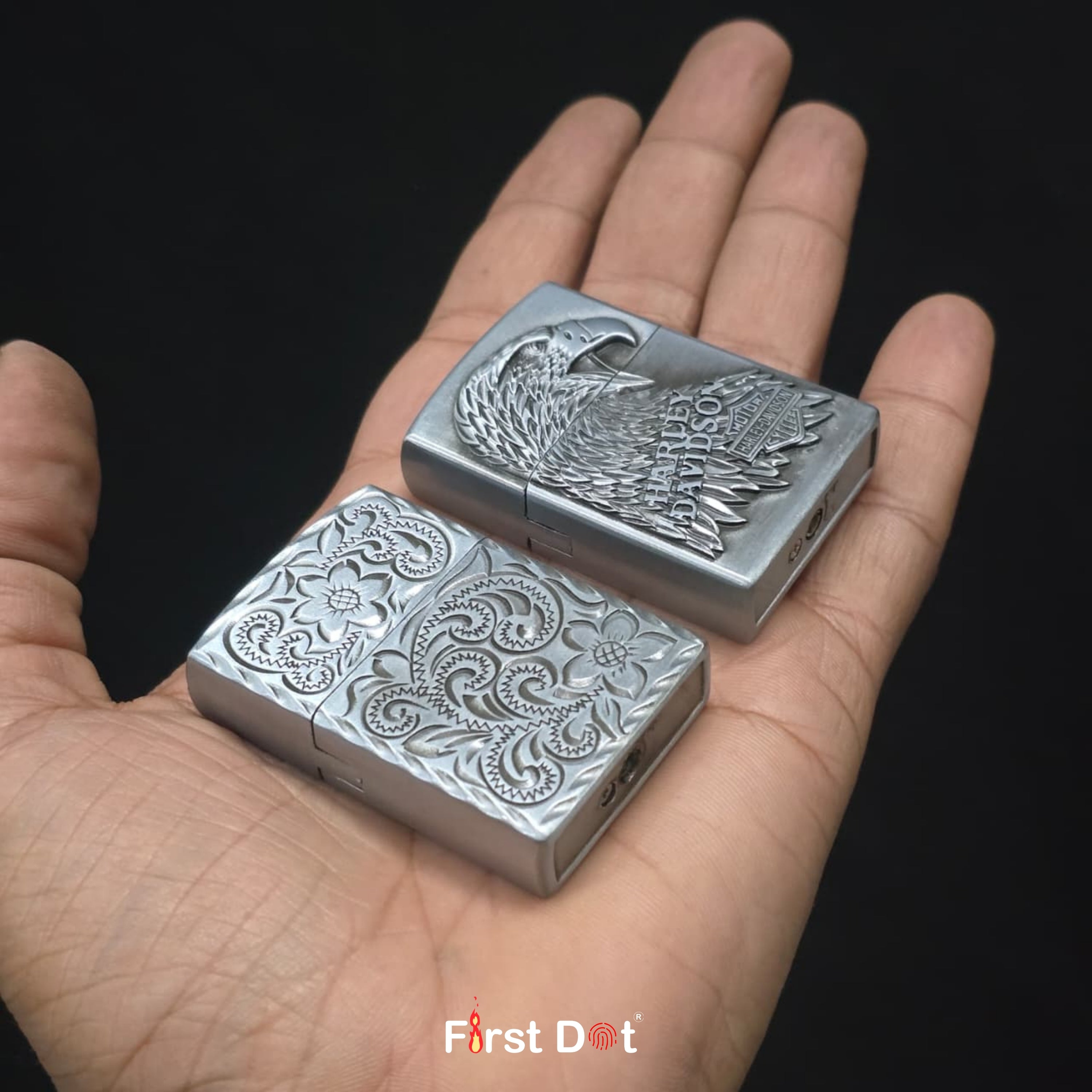 Two silver lighter with intricate designs held in a hand against a black background, branded 'First Dot'.