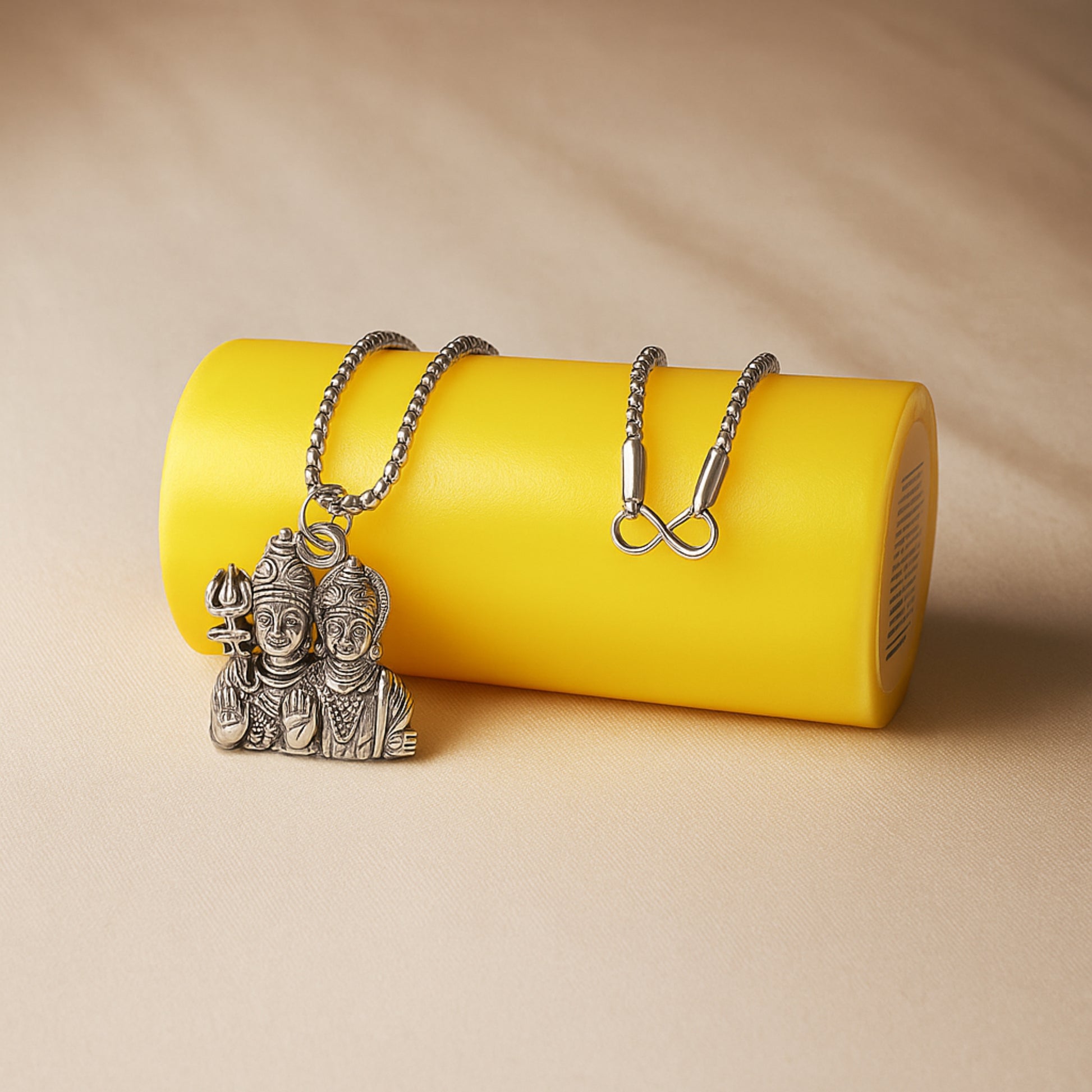 Two silver necklaces with unique pendants on a yellow cylindrical object against a beige background ShivaParvatiLocketwithChain