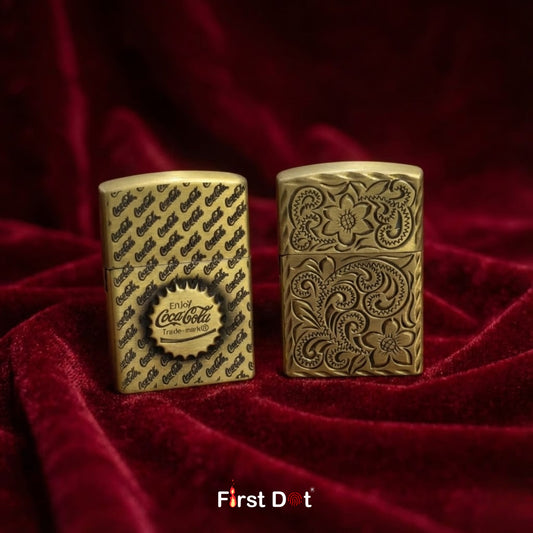 Two gold lighters with intricate designs on a red fabric background, featuring Coca-Cola branding.