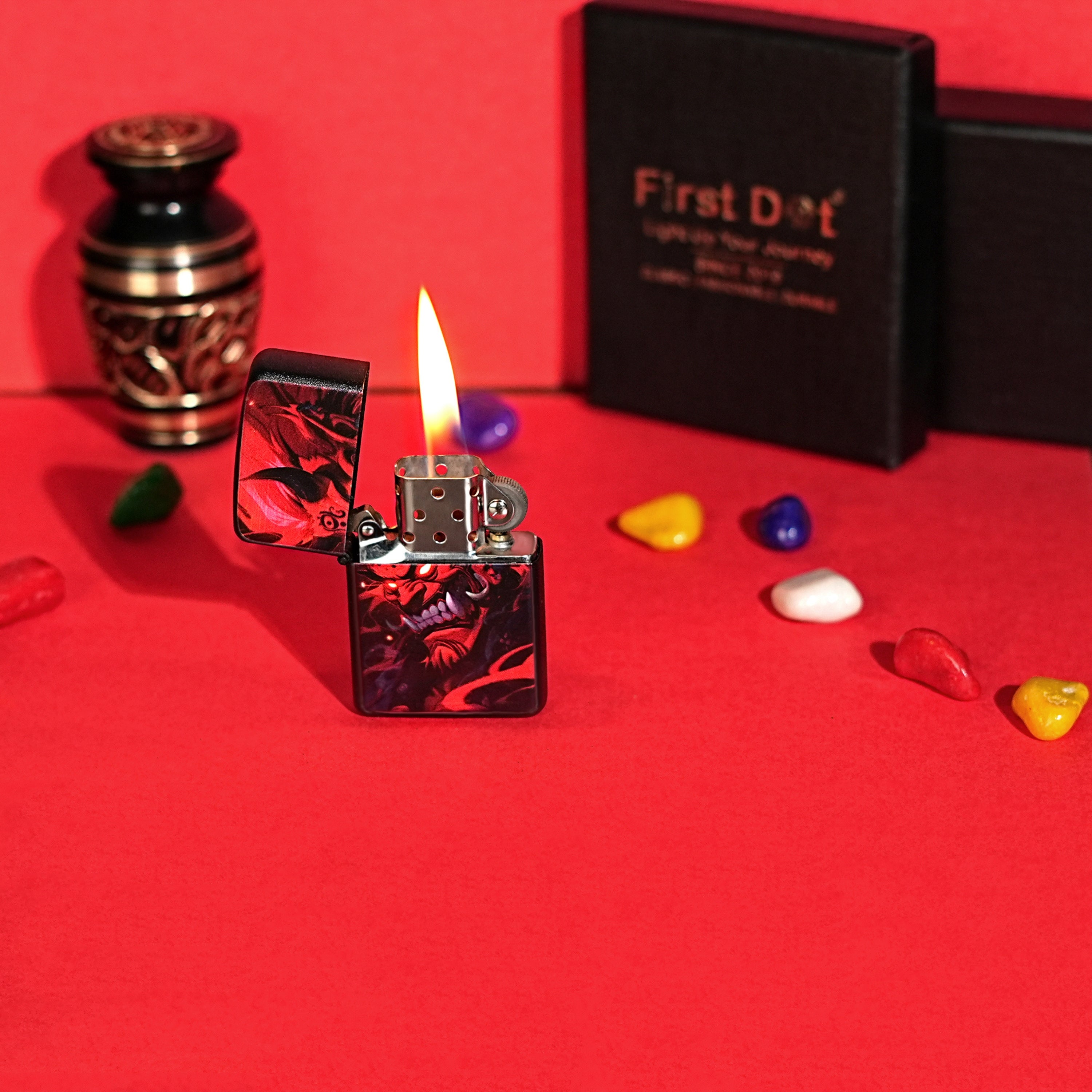 Lighter with flame on a red surface, surrounded by decorative stones and a black box labeled 'First Dot'. Hannya Demon Premium Refillable Cigarette Lighter