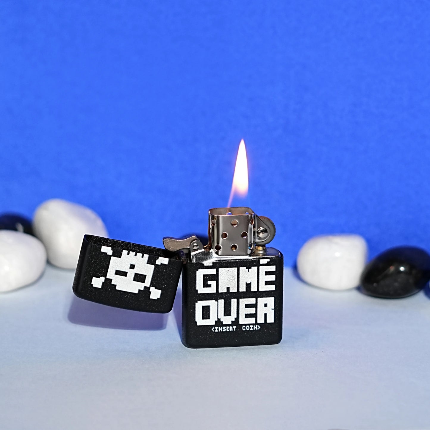 Black lighter with 'Game Over' text and pixelated design, lit on a blue background with pebbles. Game Over Premium Refillable Cigarette Lighter