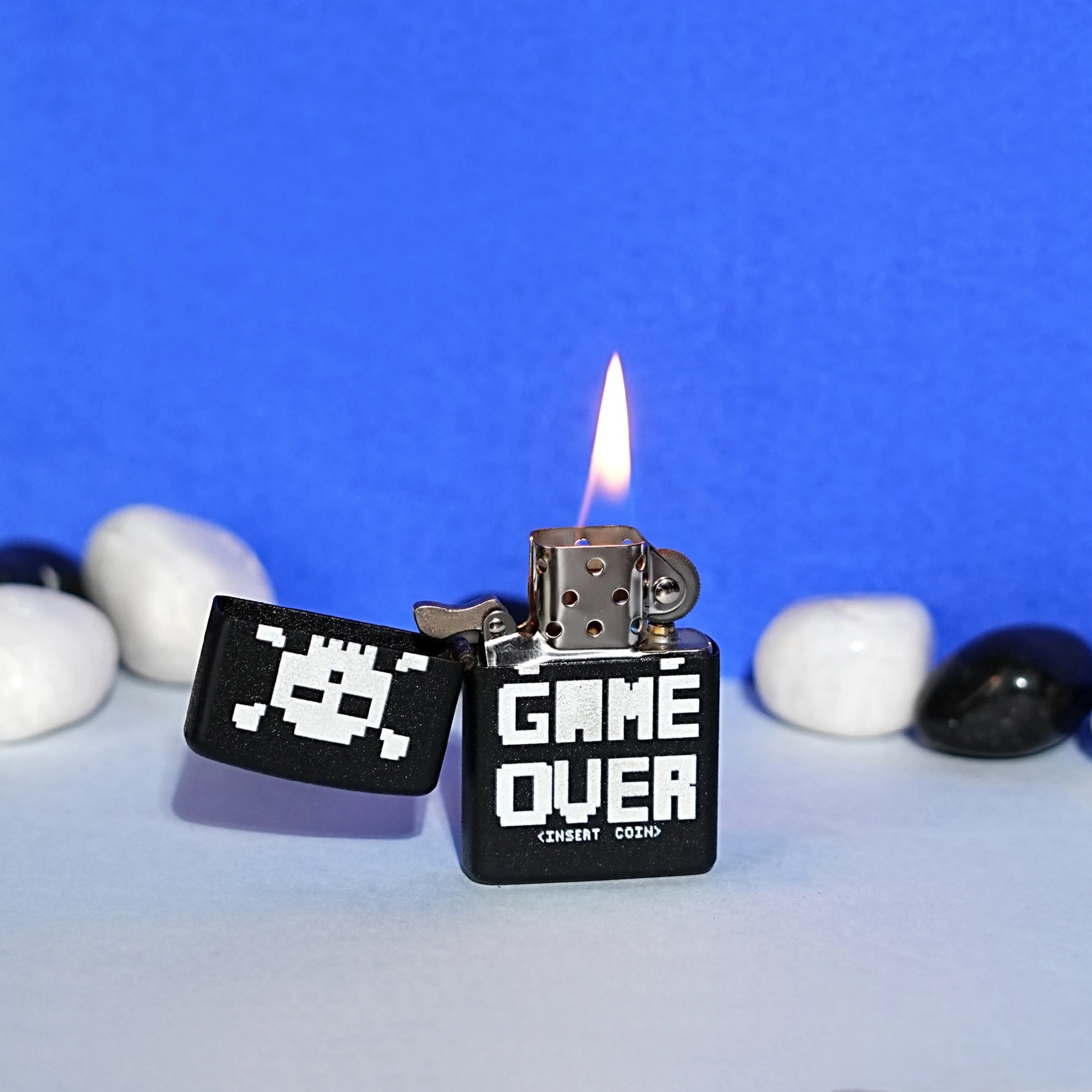 Black lighter with 'Game Over' text and pixelated design, lit on a blue background with pebbles. Game Over Premium Refillable Cigarette Lighter